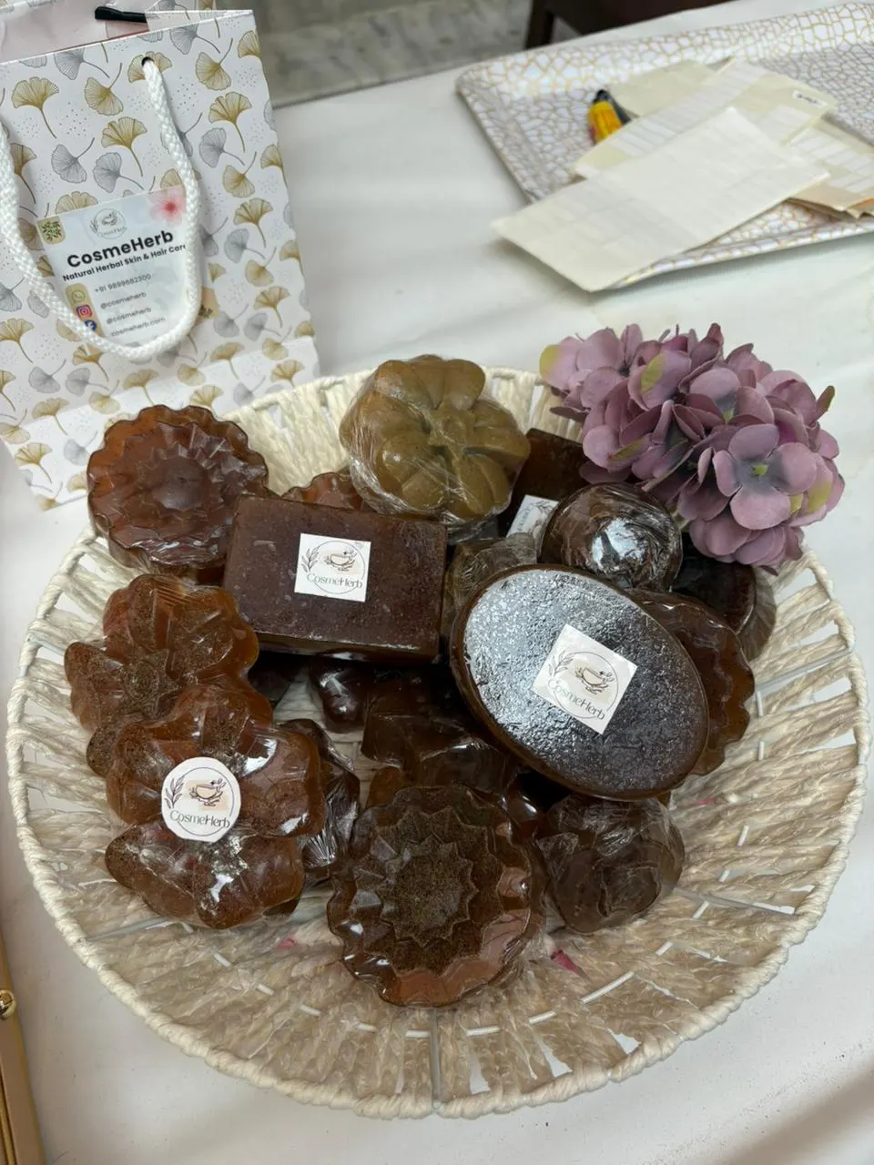 A woven basket of wrapped, moulded herbal soaps with Cosmeherb stickers, next to a printed Cosmeherb gift bag.